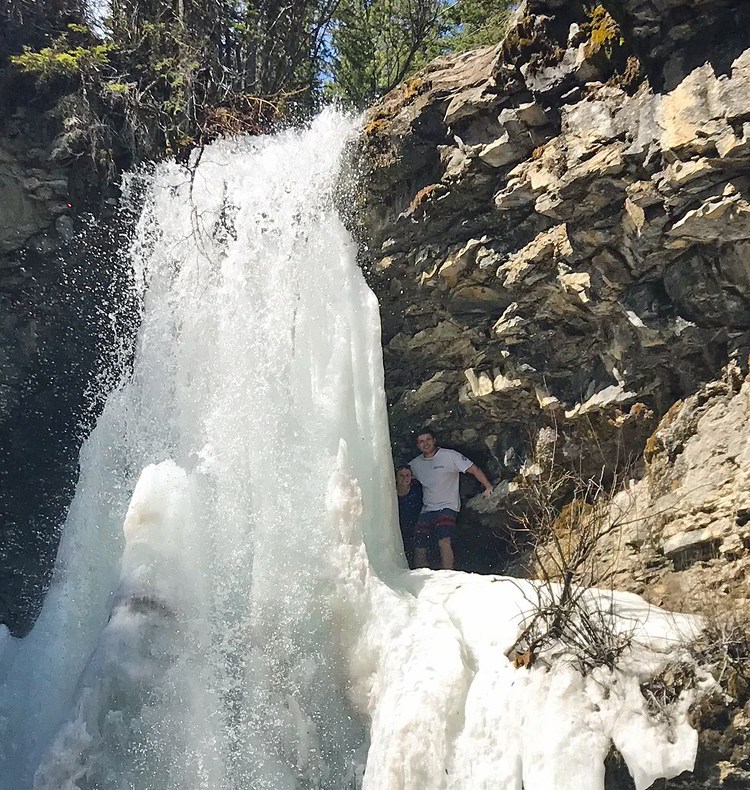 An image of a couple peaking out from behind Troll Falls in Kananaskis, Alberta