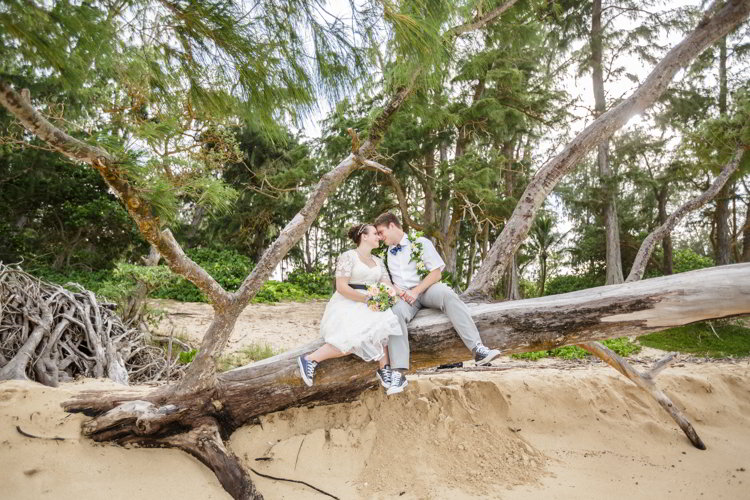 An image of a bride and groom sitting on a log on a beach in Hawaii