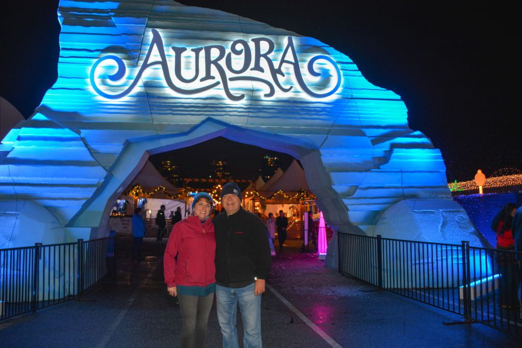 An image of a couple standing in front of the sign for the Aurora Winter Festival - Vancouver Christmas Lights