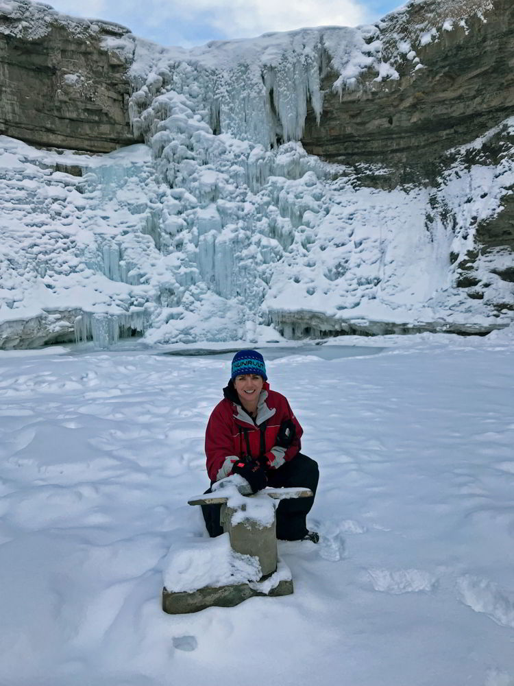 An image of a woman crouched in front of the frozen Crescent Falls - Abraham Lake, Alberta
