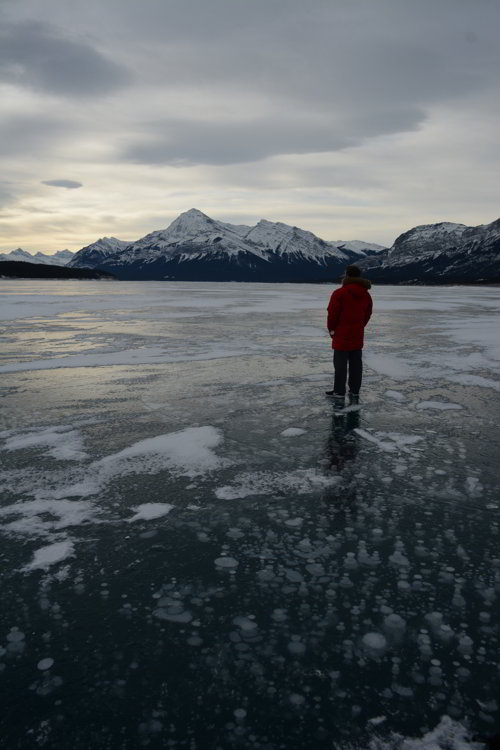 Image of a person standing on frozen Abraham Lake with methane bubbles trapped in the ice and a mountain in the background - Avoid the Winter Blues.
