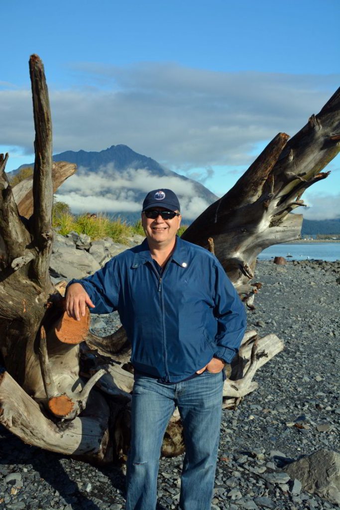 An image of a man standing in front of a large piece of driftwood on the shore of Resurrection Bay in Seward, Alaska USA 