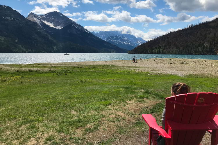 An image of a young woman sitting in a red chair by Upper Waterton Lake in Alberta, Canada. 
