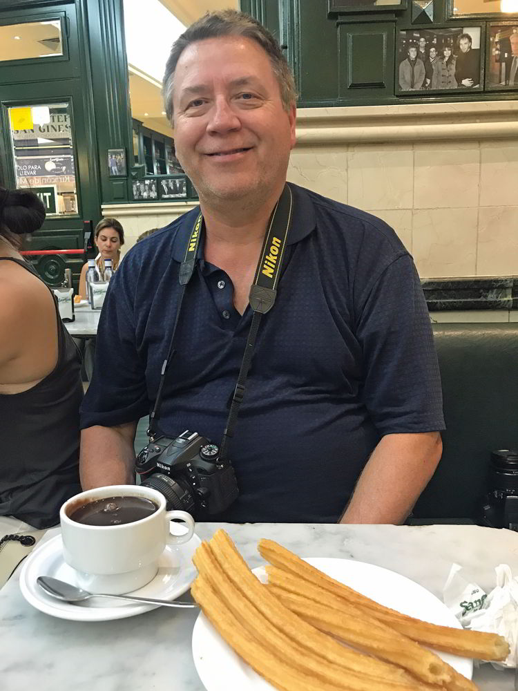 An image of a man sitting in front of a plate of churros and a cu pof hot chocolate in Madrid, Spain. 