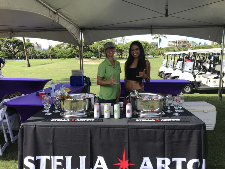 An image of two women handing out drinks at Roy's Golf Classic at the Hawaii Food and Wine Festival in Maui. 