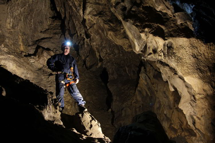 An image of the inside of the Rat's Nest Cave and a man standing on a rock with a helmet and head lamp on - Canmore Cave Tours