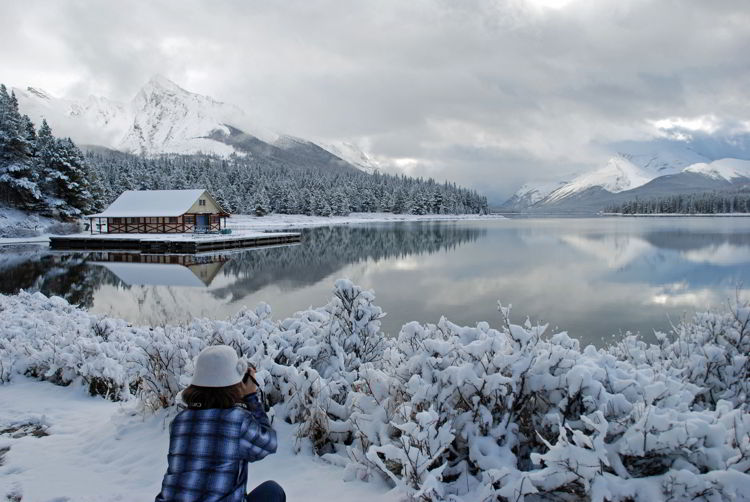 An image of a woman taking a picture of snowy Maligne Lake in Jasper in winter. 
