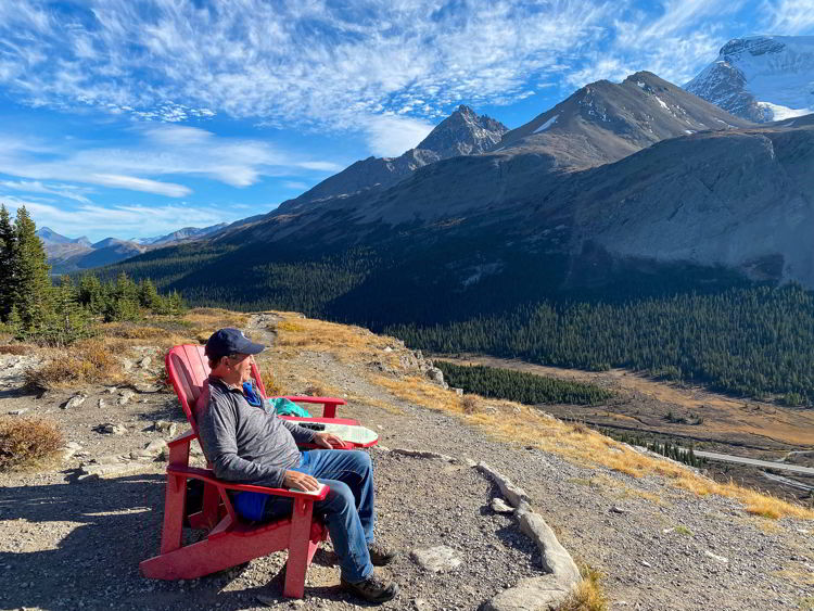 An image of a man sitting in a Parks Canada red chair taking in the view of the Athabasca Glacier as seen from the Wilcox Pass trail on the Icefields Parkway in Jasper National Park in Alberta, Canada. 