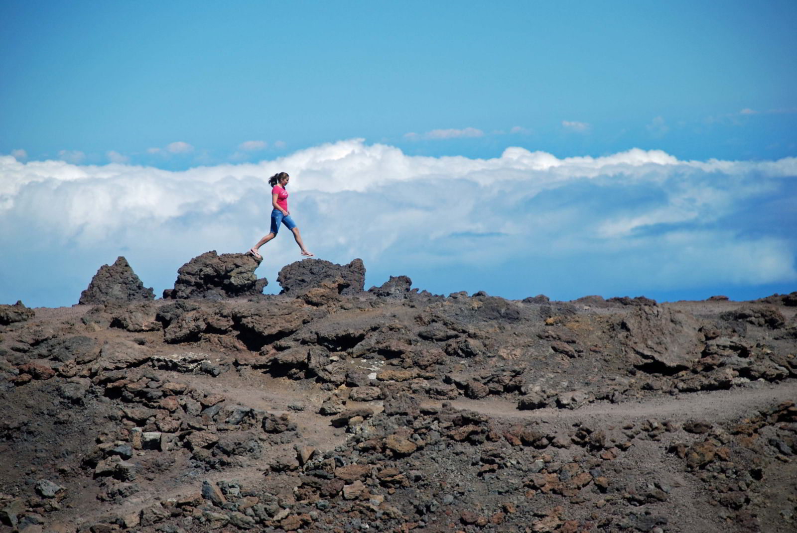 An image of a hiker jumping from one rock to the next with clouds behind them on Haleakala on the island of Maui, Hawaii - Hiking Maui